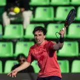 Giocatore di padel in azione, con la racchetta sollevata e la palla in volo. Le tribune verdi sullo sfondo creano un contrasto vibrante.