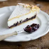 Una fetta di crostata ricotta e visciole, cosparsa di zucchero a velo, su un piatto bianco. Un cucchiaino pieno di visciole confettate accanto.