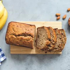 Un delizioso banana bread appena sfornato, affettato su un tagliere di legno, con banane mature e mandorle sparse.