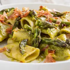 Paccheri conditi con una cremosa salsa verde e asparagi selvatici croccanti, guarniti con scaglie di prosciutto.