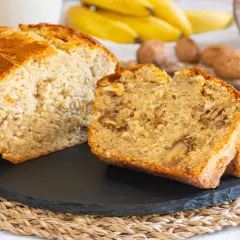 Fetta di banana bread senza burro, soffice e ricca di noci, con banane mature e noci sullo sfondo.