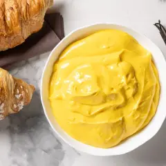 Una ciotola di cremosa crema pasticcera senza glutine, con accanto due croissant e un baccello di vaniglia.