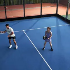 Coppia gioca a padel in campo blu. Lui colpisce la palla, lei attende il suo turno. Si può giocare a padel in 2.