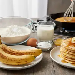 Ingredienti per pancake alla banana: banane mature, farina, uova, latte, zucchero e miele. Una pila di pancake dorati guarniti con banane e sciroppo.