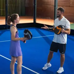 Coppia sorridente si prepara per una partita di padel misto, racchette in mano sul campo blu.