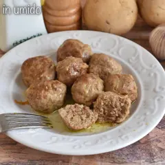 Polpette in umido succulente su un piatto bianco, con patate e aglio sullo sfondo. Una forchetta è pronta per gustarle.