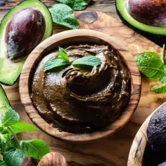 Una ciotola di legno contiene una deliziosa crema di avocado, guarnita con foglie di menta fresca. Avocadi maturi e foglie verdi circondano la ciotola.