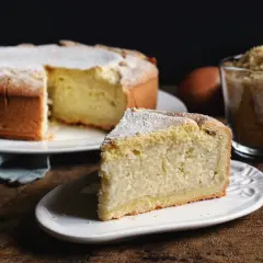 Una fetta di torta di riso, soffice e cosparsa di zucchero a velo, accanto alla torta intera e a una tazza di riso crudo.