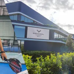 Rafa Nadal sorride su un campo da padel, con la sua accademia sullo sfondo. Un campione che ispira nel mondo del nadal padel.