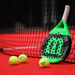 Racchetta da padel verde e nera appoggiata alla rete, con palline gialle sul campo rosso. Quanto dura una racchetta da padel? Dipende dall'uso!