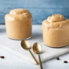 Due bicchieri di cremosa schiuma di caffè, perfetti per una ricetta crema di caffè senza panna. Due cucchiaini dorati e chicchi di caffè completano la scena.