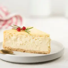 Una fetta di cheesecake con yogurt greco, guarnita con rosmarino e bacche rosse, su un piatto bianco con cucchiaino dorato.