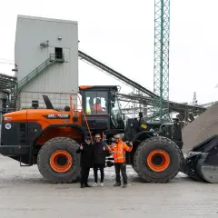 Un imponente **taipan nero** (pale caricatrici) domina la scena in un impianto di lavorazione inerti. Tre uomini sorridono, pronti per il lavoro.