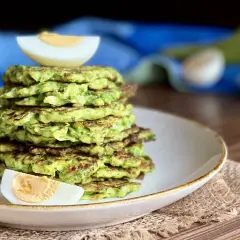 Una pila di rosti di zucchine dorati, guarniti con spicchi di uovo sodo, su un piatto bianco.