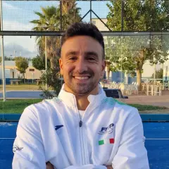 Aris Patiniotis sorride con le braccia conserte su un campo da padel blu. Indossa una giacca bianca con logo e bandiera italiana.