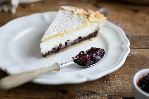 Una fetta di crostata ricotta e visciole, cosparsa di zucchero a velo, su un piatto bianco. Un cucchiaino pieno di visciole confettate accanto.