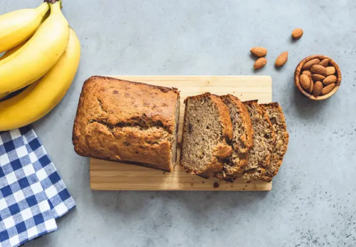 Un delizioso banana bread appena sfornato, affettato su un tagliere di legno, con banane mature e mandorle sparse.