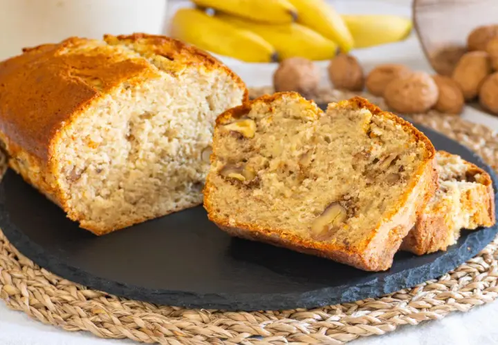 Fetta di banana bread senza burro, soffice e ricca di noci, con banane mature e noci sullo sfondo.