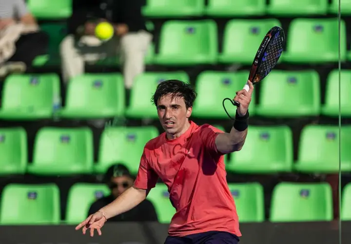 Giocatore di padel in azione, con la racchetta sollevata e la palla in volo. Le tribune verdi sullo sfondo creano un contrasto vibrante.