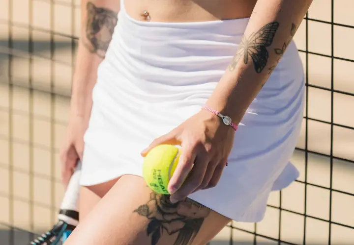 Braccialetto con ciondolo racchetta da padel, tatuaggio farfalla e rosa, palla da padel in mano.