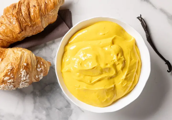 Una ciotola di cremosa crema pasticcera senza glutine, con accanto due croissant e un baccello di vaniglia.