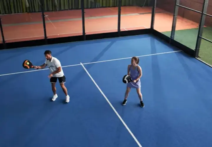 Coppia gioca a padel in campo blu. Lui colpisce la palla, lei attende il suo turno. Si può giocare a padel in 2.