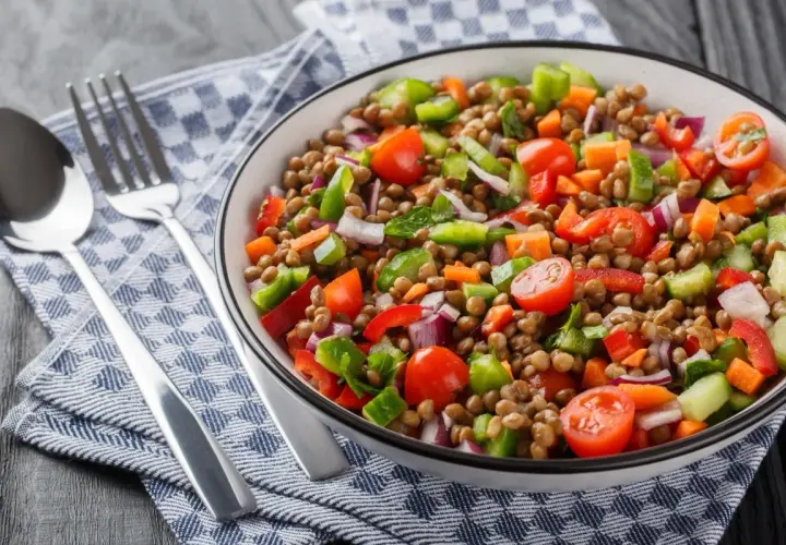 Un'insalata di lenticchie colorata e fresca, con pomodorini, peperoni, cipolla rossa e carote. Servita in una ciotola con posate su un tovagliolo a quadri.