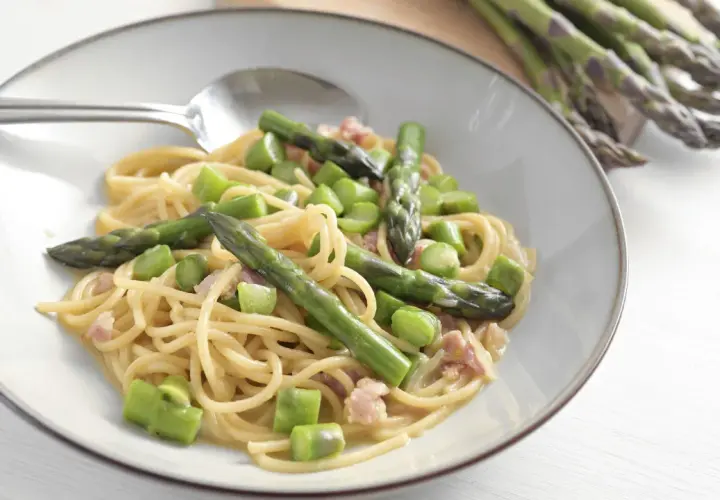 Un piatto di spaghetti con asparagi freschi e cubetti di speck croccante, condito con una cremosa salsa.
