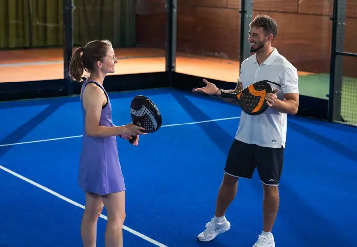 Coppia sorridente si prepara per una partita di padel misto, racchette in mano sul campo blu.