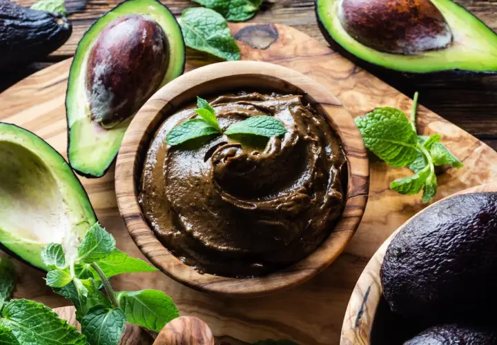 Una ciotola di legno contiene una deliziosa crema di avocado, guarnita con foglie di menta fresca. Avocadi maturi e foglie verdi circondano la ciotola.