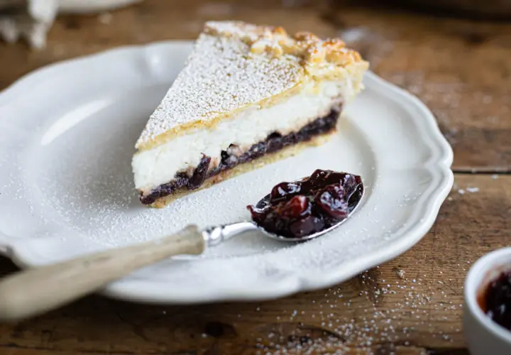 Una fetta di crostata ricotta e visciole, cosparsa di zucchero a velo, su un piatto bianco. Un cucchiaino pieno di visciole confettate accanto.