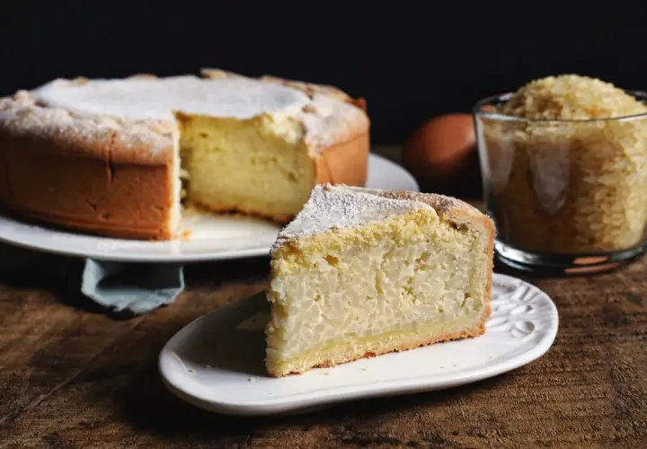 Una fetta di torta di riso, soffice e cosparsa di zucchero a velo, accanto alla torta intera e a una tazza di riso crudo.
