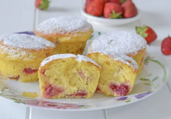 Morbidi muffin alle fragole, cosparsi di zucchero a velo, su un piatto fiorito. Uno è tagliato a metà, rivelando il ripieno succoso.