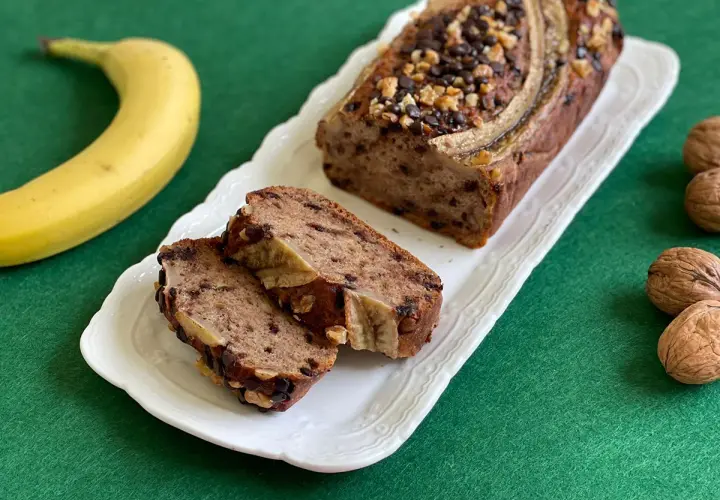 Delizioso banana bread light con gocce di cioccolato e noci, guarnito con banana e servito su un piatto bianco.