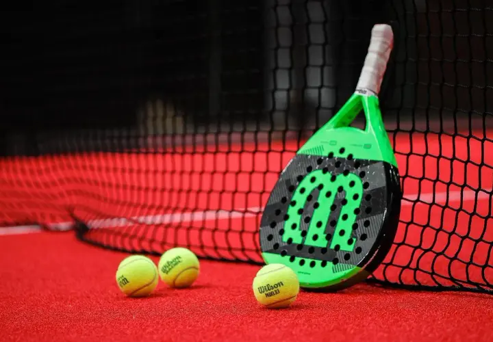 Racchetta da padel verde e nera appoggiata alla rete, con palline gialle sul campo rosso. Quanto dura una racchetta da padel? Dipende dall'uso!