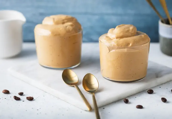 Due bicchieri di cremosa schiuma di caffè, perfetti per una ricetta crema di caffè senza panna. Due cucchiaini dorati e chicchi di caffè completano la scena.