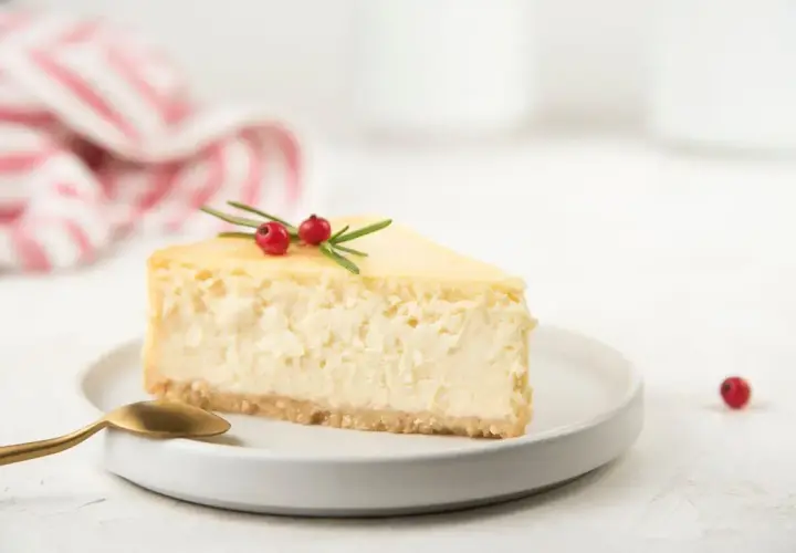 Una fetta di cheesecake con yogurt greco, guarnita con rosmarino e bacche rosse, su un piatto bianco con cucchiaino dorato.