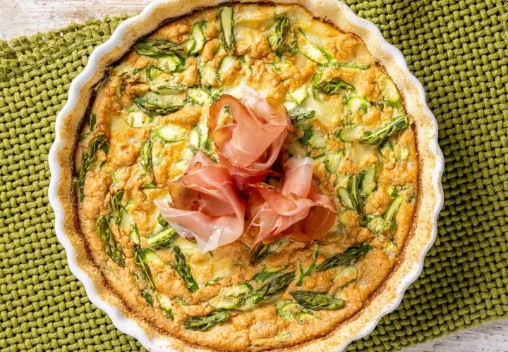 Frittata di asparagi al forno, dorata e invitante, guarnita con fette di prosciutto crudo. Un piatto perfetto per un pranzo leggero.
