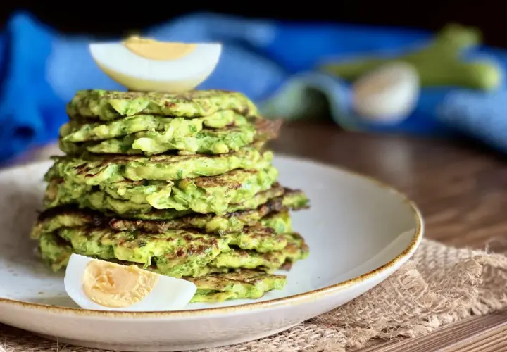Una pila di rosti di zucchine dorati, guarniti con spicchi di uovo sodo, su un piatto bianco.