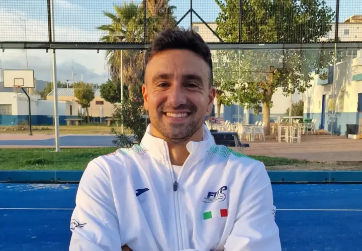 Aris Patiniotis sorride con le braccia conserte su un campo da padel blu. Indossa una giacca bianca con logo e bandiera italiana.