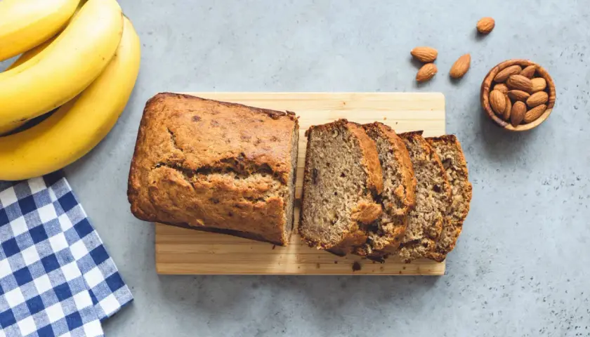 Un delizioso banana bread appena sfornato, affettato su un tagliere di legno, con banane mature e mandorle sparse.