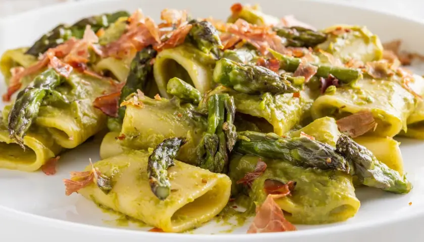 Paccheri conditi con una cremosa salsa verde e asparagi selvatici croccanti, guarniti con scaglie di prosciutto.