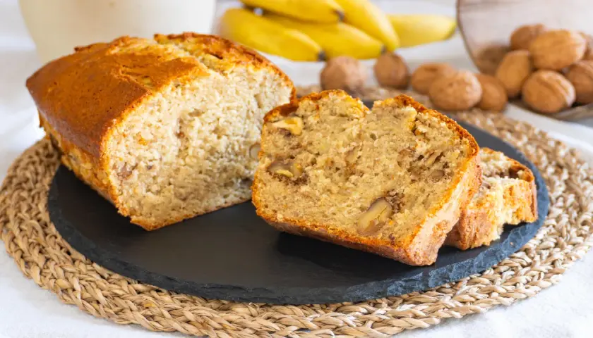 Fetta di banana bread senza burro, soffice e ricca di noci, con banane mature e noci sullo sfondo.