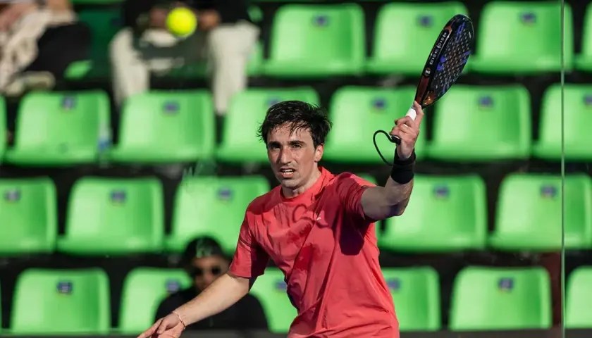 Giocatore di padel in azione, con la racchetta sollevata e la palla in volo. Le tribune verdi sullo sfondo creano un contrasto vibrante.