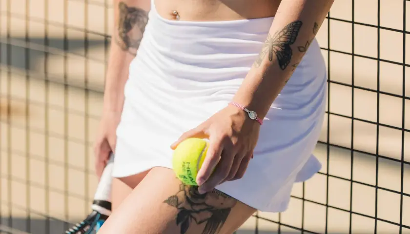 Braccialetto con ciondolo racchetta da padel, tatuaggio farfalla e rosa, palla da padel in mano.