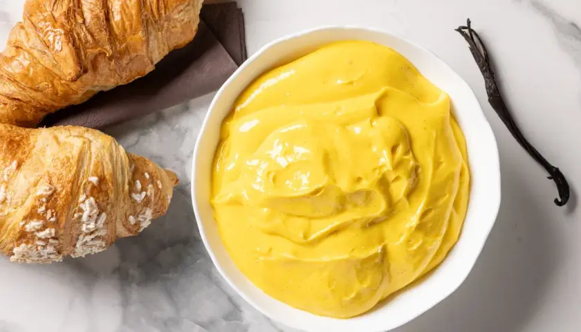 Una ciotola di cremosa crema pasticcera senza glutine, con accanto due croissant e un baccello di vaniglia.