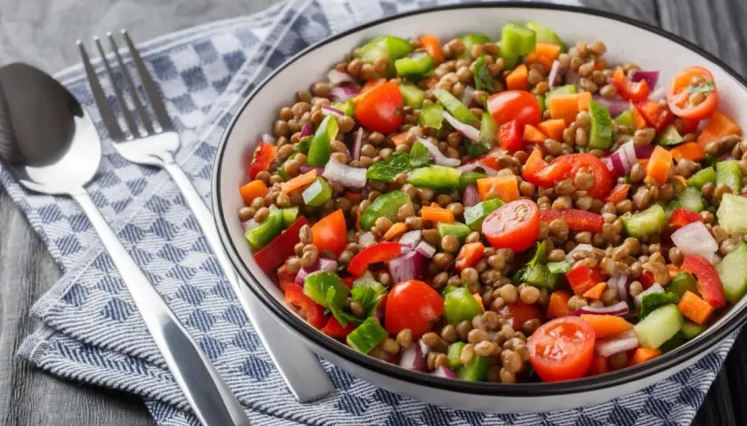 Un'insalata di lenticchie colorata e fresca, con pomodorini, peperoni, cipolla rossa e carote. Servita in una ciotola con posate su un tovagliolo a quadri.