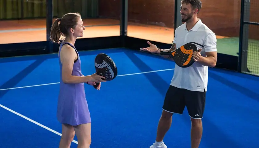 Coppia sorridente si prepara per una partita di padel misto, racchette in mano sul campo blu.