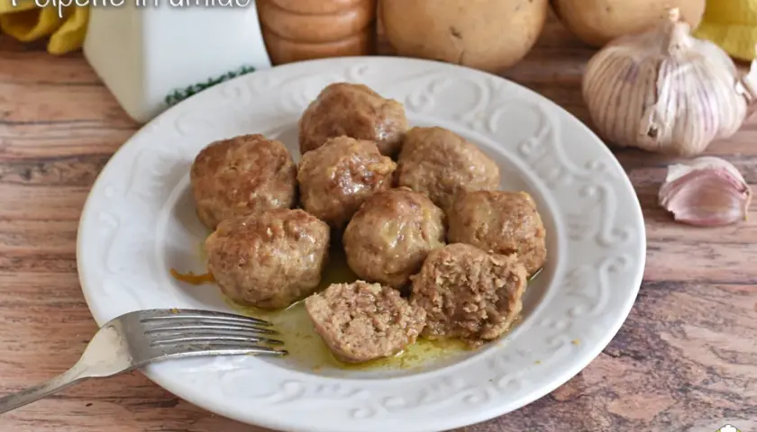 Polpette in umido succulente su un piatto bianco, con patate e aglio sullo sfondo. Una forchetta è pronta per gustarle.