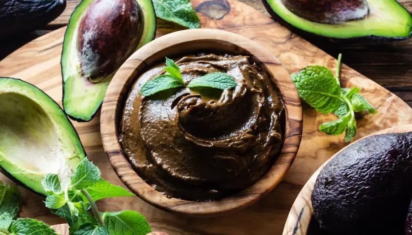 Una ciotola di legno contiene una deliziosa crema di avocado, guarnita con foglie di menta fresca. Avocadi maturi e foglie verdi circondano la ciotola.